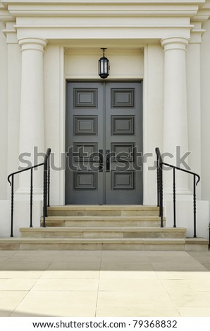 Double doors with rectangular inset patterns stand at the top of steps. There are handrails bordering the steps leading to the doors. Vertical shot.