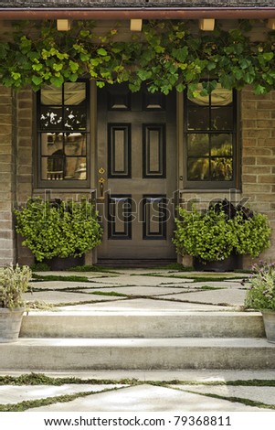 A diamond pattern concrete walkway leads to the front door of a brick home. There are potted and hanging plants around the door. Vertical shot.