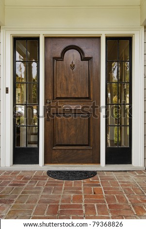 The wooden front door of a home with glass panels to each side and a brick porch. The glass is reflecting the homes opposite the door. Vertical shot.