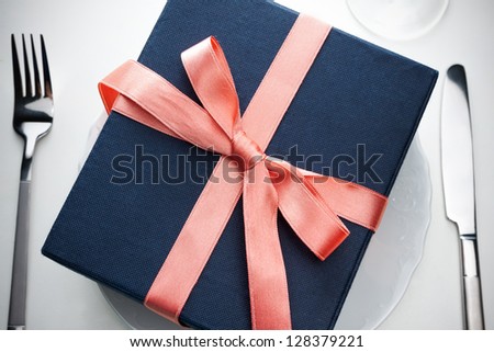 Romantic gift on a white plate in a place setting with silver cutlery wrapped in black wrapping with a decorative red bow