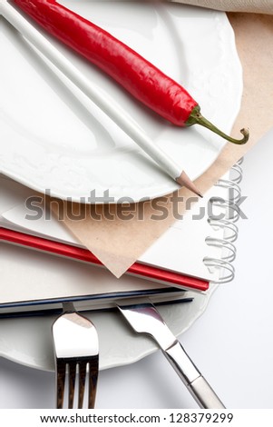 Close-up of white plates with books sandwiched in between them. A knife and fork is sticking out of one while a white pencil and red chili sits on the other.