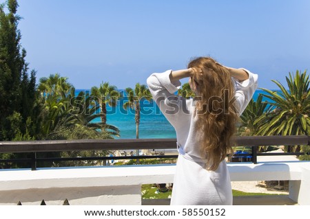 Young woman wearing white robe stands on the white hotel\'s balcony looks at the sea holding hands up to the head