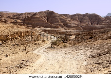 A long dusty road winds through the isolated and remote hills and valleys of the Negev desert. In the background, hills of sedimentary sandstone loom over the road.