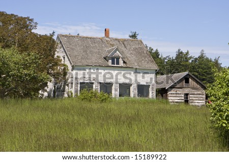 An old family farmhouse on Waldron Island, Washington viewed from across a grassy meadow. A barn stands to the right. Both of the wood buildings are very weathered.