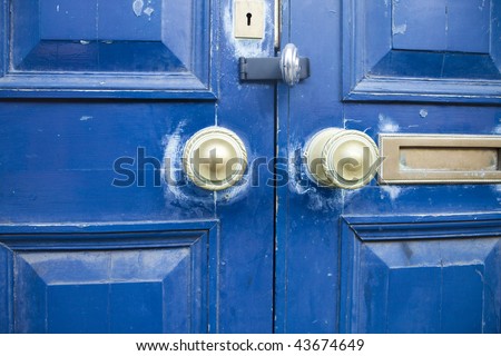 A blue door with bronze doorknobs, mailslot, and keyhole. The scuffed and faded paint together with the slightly askew door panels has a grungy feel to it.