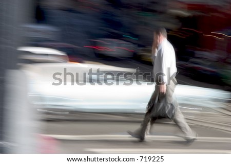 A business man walks across the street briskly while talking on a cell phone.  An intensionally long shutter gives a blurred sense of urgency to the scene.