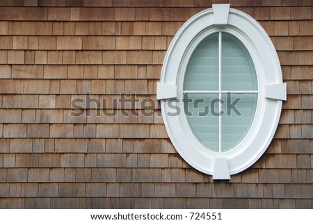 An oval window on the side of a house with shingles.