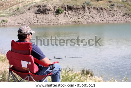 Man sitting in a red chair fishing near the edge of a pond.