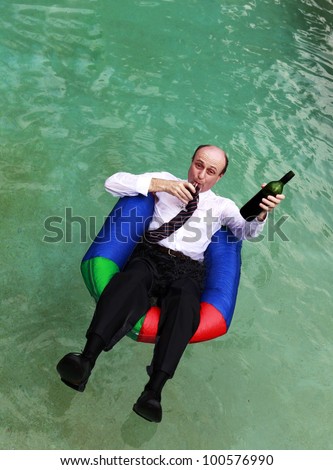 A business man dressed in business attire in a swimming pool, floating with one arm outstretched with a bottle of red wine in the outstretched arm and glass red wine in one hand.