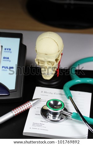 A doctorÃ¢Â?Â?s desk showing a green stethoscope and pen,  resting on a sick certificate pad,  with the other doctors tools of the trade on desk showing a blood pressure machine  and a human skull model.