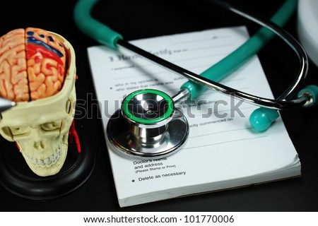 A doctorÃ¢Â?Â?s desk showing a green stethoscope and pen,  resting on a sick certificate pad,  with the doctors tools of the trade on desk including a blood pressure machine and model of a human skull.
