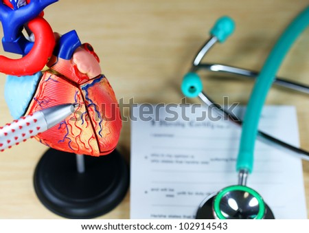 A doctorÃ¢Â?Â?s desk showing a green stethoscope, on a sick certificate pad in the background, and in the foreground the doctors pen point to a model of the heart, indicating where the patients problem is.