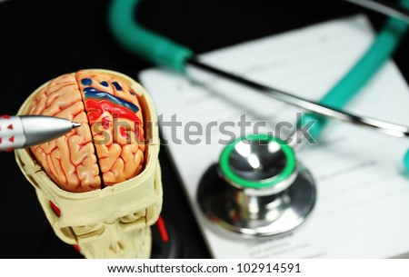 A doctorÃ¢Â?Â?s desk showing a green stethoscope and pen, on a sick certificate pad, with the other doctors tools of  and model of a human skull, with the Doctors pen pointing to where the problem is.