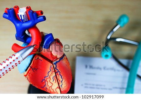 A doctorÃ¢Â?Â?s desk showing a green stethoscope on a sick certificate pad in the background, and in the foreground the doctors pen point to a model of the heart, indicating where the patients problem is.