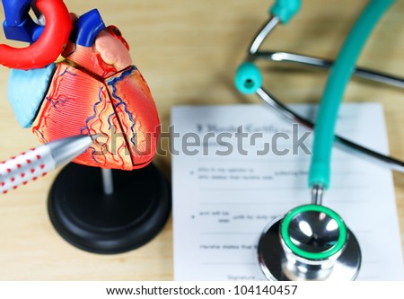 A doctorÃ¢Â?Â?s desk showing a green stethoscope, resting on a sick certificate pad in the background, and in the foreground the doctorÃ¢Â?Â?s pen point to a model of the heart, indicating patientÃ¢Â?Â?s problem is.