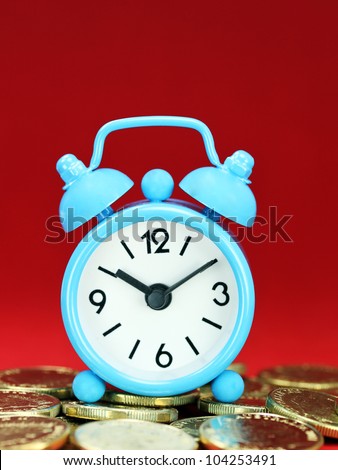 A light blue alarm clock placed on some golden coins with a red background, asking the question how long before your investment matures?