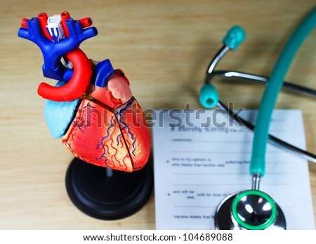 A doctorÃ¢Â?Â?s desk showing a green stethoscope, resting on a sick certificate pad in the background, and in the foreground a detailed model of the human heart.