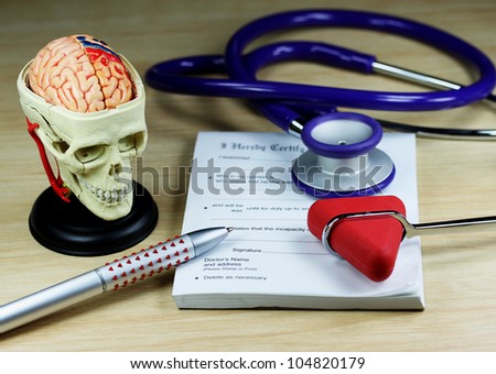 A doctorÃ¢Â?Â?s desk showing a purpe stethoscope and pen, resting on a sick certificate pad, with the other doctorÃ¢Â?Â?s tools of the trade on desk including a model of a human skull and a patella hammer.