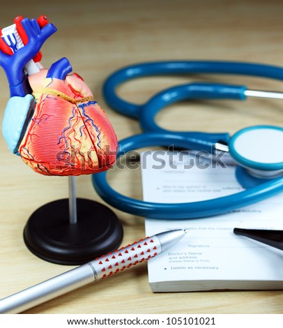 A doctors desk showing a green stethoscope, resting on a sick certificate pad in the background, and in the foreground a detailed model of the human heart.