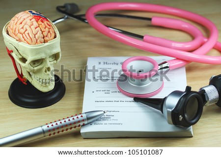 A doctors desk showing a green stethoscope and pen, resting on a sick certificate pad, with the other doctors tools of the trade on desk including a model of a human skull, with the brain exposed.
