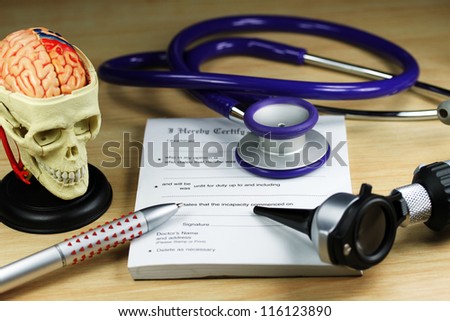 A doctorÃ¢Â?Â?s desk showing a purple stethoscope and pen, resting on a sick certificate pad, with the other doctorÃ¢Â?Â?s tools of the trade on desk including a model of a human skull, with the brain exposed.