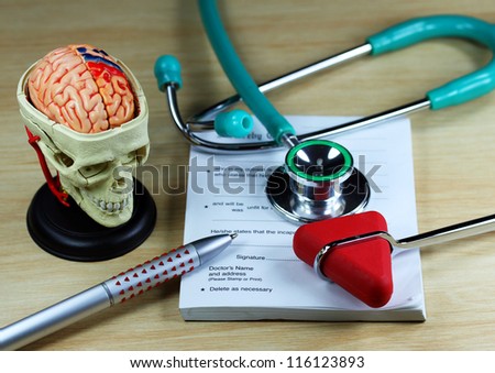 A doctorÃ¢Â?Â?s desk showing an aqua stethoscope and pen, resting on a sick certificate pad, with the other doctorÃ¢Â?Â?s tools of the trade on desk including a model of a human skull, with the brain exposed.
