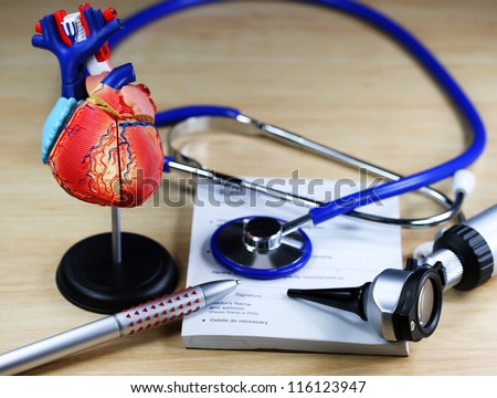 A doctorÃ¢Â?Â?s desk showing a blue stethoscope, resting on a sick certificate pad with the other diagnostic doctors tools and a detailed model of the human heart.