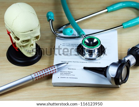 A doctorÃ¢Â?Â?s desk showing a green stethoscope and pen, resting on a sick certificate pad, with the other doctorÃ¢Â?Â?s tools of the trade on desk including a model of a human skull.