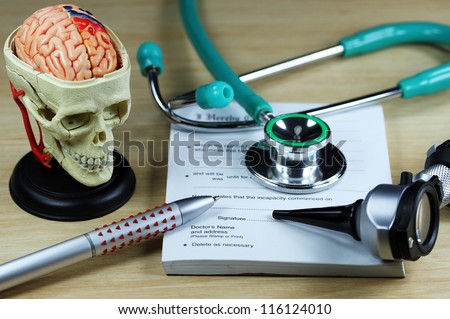 A doctorÃ¢Â?Â?s desk showing a green stethoscope and pen, resting on a sick certificate pad, with the other doctorÃ¢Â?Â?s tools of the trade on desk including a model of a human skull, with the brain exposed.