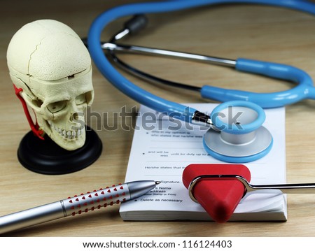A doctorÃ¢Â?Â?s desk showing a purple stethoscope, patella hammer and pen, resting on a sick certificate pad, with the other doctorÃ¢Â?Â?s tools of the trade on desk including a model of a human skull.