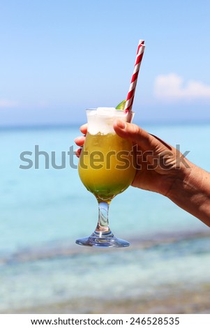 A beautiful yellow cocktail held in a ladies hand with the tropical aqua ocean and blue skein the back ground