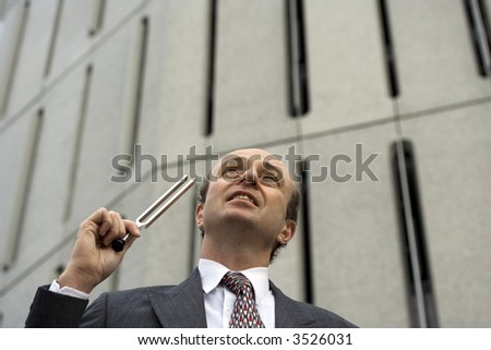 Businessman outside his office in the Central Business District, with a tuning fork held to his ear, indicating are you in tune with your business or its needs?