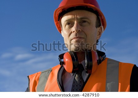 Engineer looking thoughtful, with a beautiful blue sky behind him, wearing a red safety helmet, high visibility orange vest over his suit and wearing ear muffs around his neck
