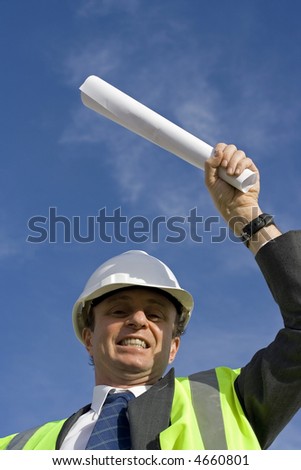 Engineer looking happy, with a beautiful blue sky behind him, wearing a red white safety helmet, wearing a yellow high visibility vest and carrying the blueprints after being successful