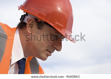 Engineer looking puzzled, with a beautiful blue sky behind him, wearing a red safety helmet, well-dressed with an orange high visibility reflective vest on