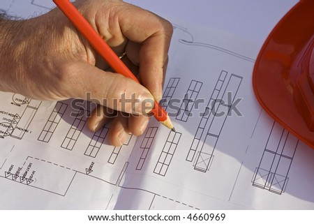 Engineers hand holding a pencil poised over the blueprints with his red safety helmet resting on the plans