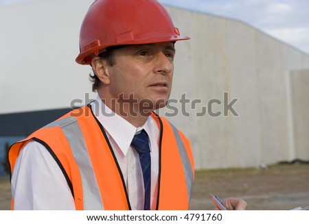 Engineer looking focussed, with a building behind him being constructed  sky behind him, wearing a red safety helmet and wearing an orange high visibility vest.