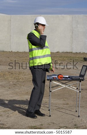 Engineer looking thoughtful on the building site, with blue sky behind him, wearing a white safety helmet, high visibility yellow vest over his suit and on a mobile telepone