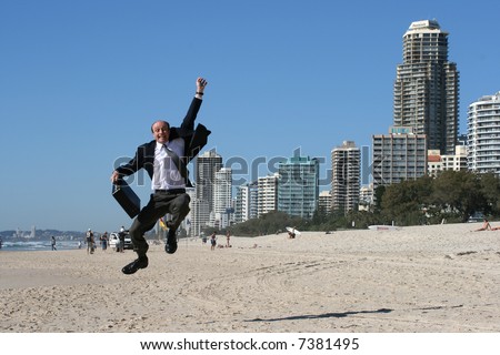 Businessman jumping for joy on a wonderful beach carrying his briefcase, wearing a suit with the high rise buildings on the shoreline.