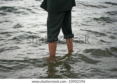 Businessman dressed in business attire wading in the sea with his trousers rolled up, testing the water.