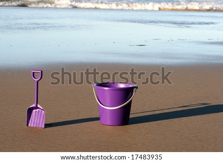 A purple plastic bucket and spade on the beach with the ocean coming in behind them