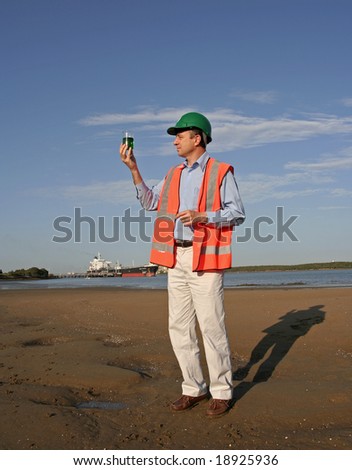 A environmental engineer on the mudflats examining a sample of  oil from the ship docked behind him, showing the estuary and beautiful blue sky, wearing orange reflective vest and green safety helmet