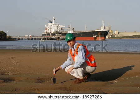 A environmental engineer on the mudflats examining a sample of oil from the ship docked behind him, showing the estuary and beautiful blue sky, wearing orange reflective vest and green safety helmet