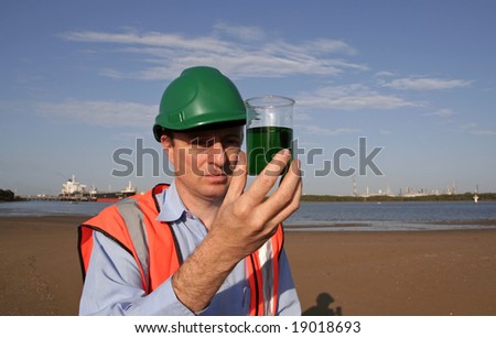 An environmental engineer on the mudflats examining a sample of oil from the ship docked behind him, showing the estuary and beautiful blue sky, wearing orange reflective vest and green safety helmet