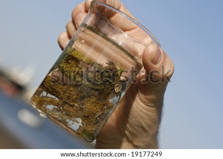 A sample taken from the ocean with a plantlike creature in a glass specimen jar, behind the sample is a tanker in the docks.