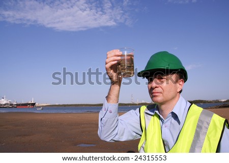 An environmental engineer on the mudflats examining an unusual  plant specimen found on the mudflats in the background is a tanker moored, wearing a yellow reflective vest and green safety helmet.