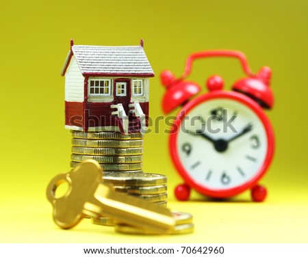 A house placed on a pile of gold coins with a golden key in front and a red alarm clock behind, asking the question is it time to buy that dream house?