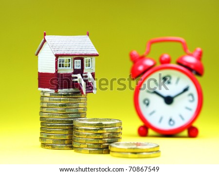 A house placed on a stack of gold coins with a red alarm clock behind, asking the question is it time to buy that dream house?
