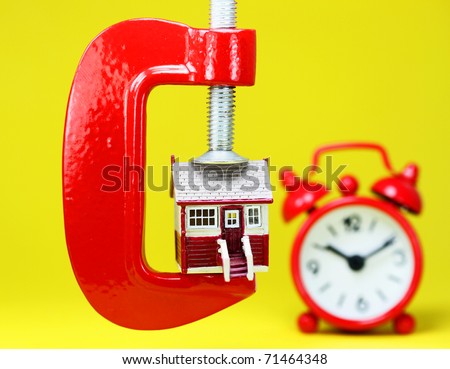 A model house placed in a red clamp with a red clock in the background indicating is it time to act upon buying a home.