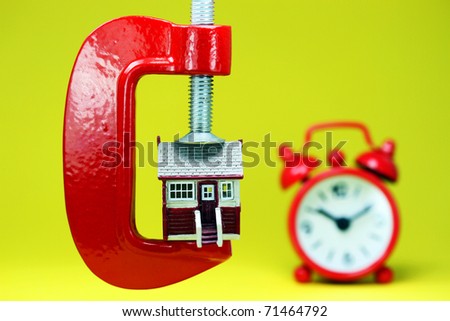 A model house placed in a red clamp with a red clock in the background indicating is it time to act upon buying a home.
